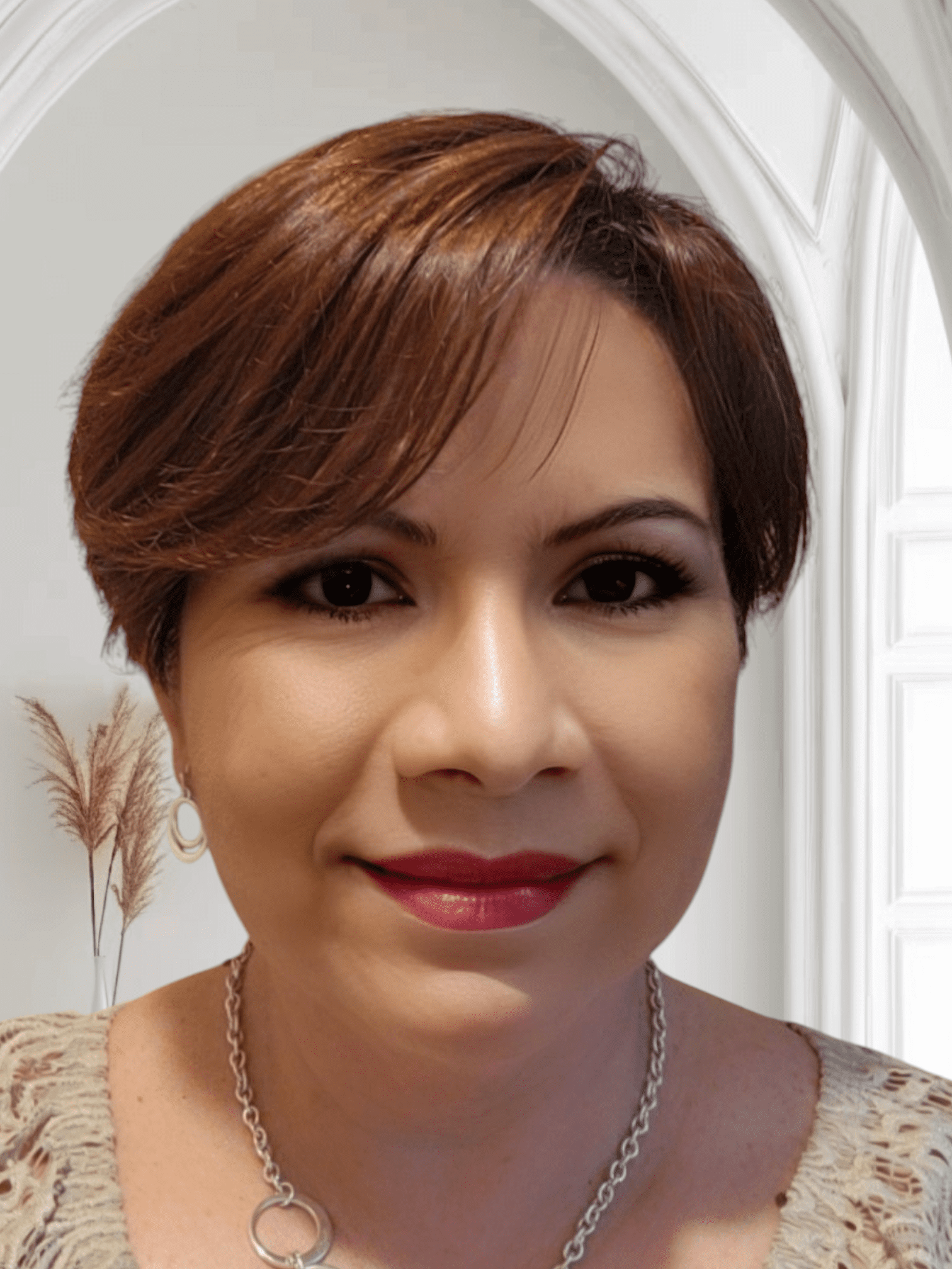 A person with short brown hair, wearing hoop earrings and a necklace, smiles in front of a white archway.